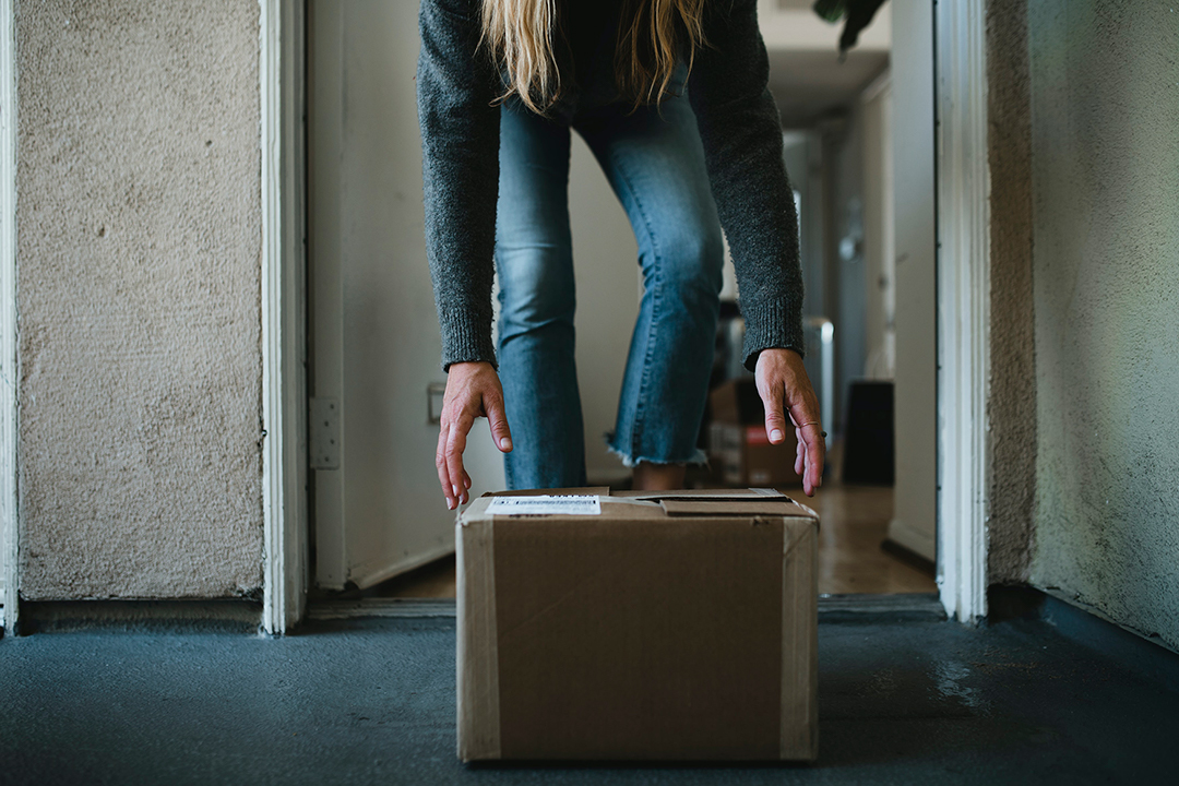 person bending down to pick up a box outside their door. Photo. 