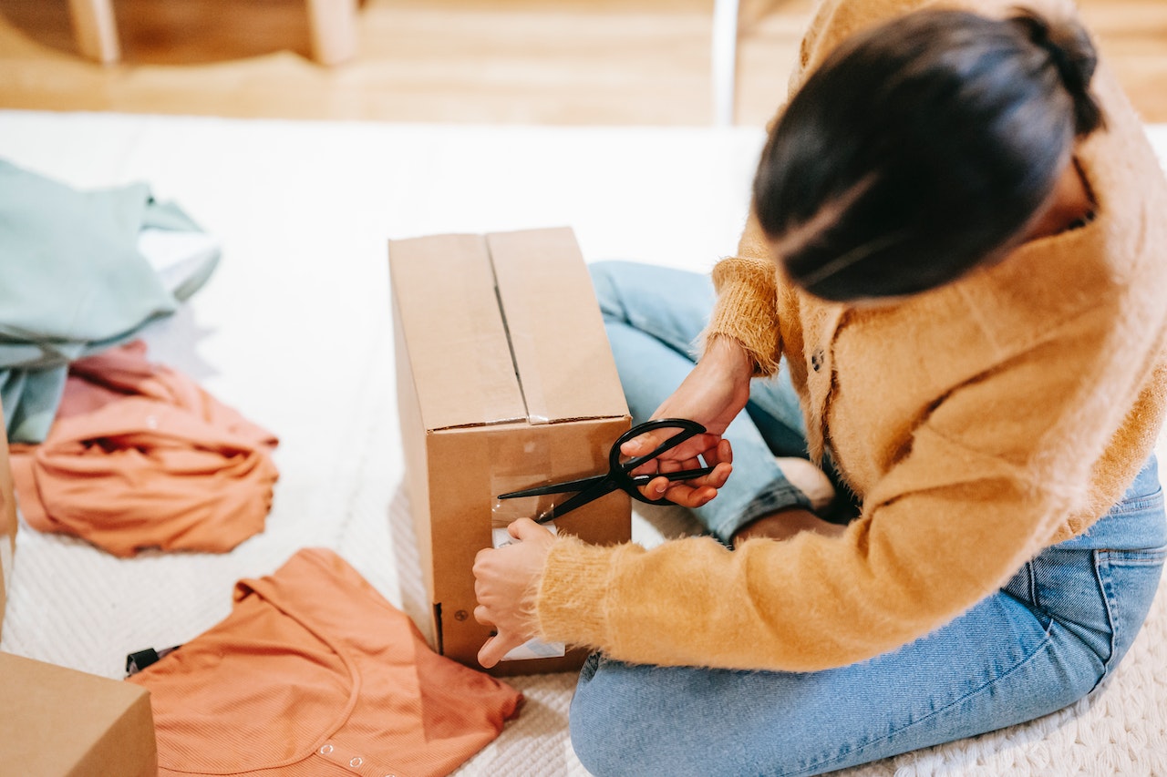 person sitting on the floor taping a box shut, surrounded by clothing items. Photo. 