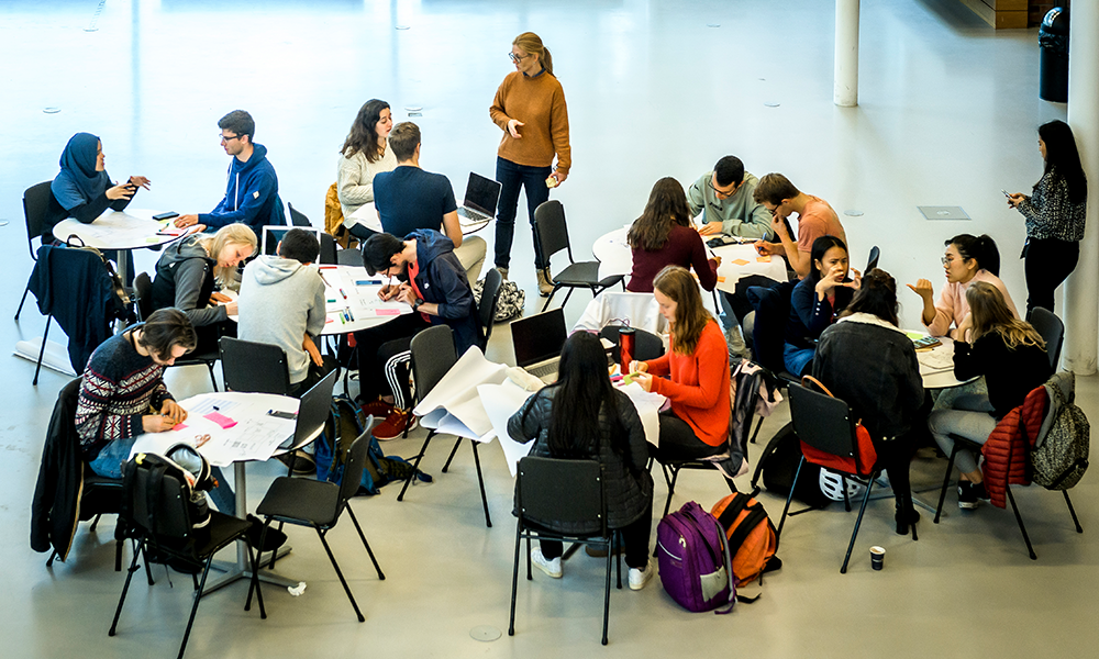 People in a room sitting around different tables working together. Photo.